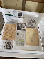 Historische Dokumente liegen auf einem Schreibtisch neben einem Laptop mit CMS-Ansicht zur Bildbearbeitung.