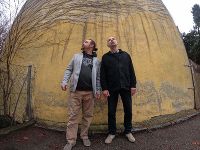 Das Bild zeigt zwei Männer, die vor einer gelben Wand stehen