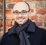 Person mit Brille und Schal lächelt in die Kamera. Im Hintergrund eine Backsteinmauer.