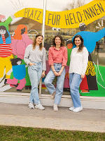 Drei junge Menschen stehen lächelnd vor einem bunten Wandbild mit dem Schriftzug "Platz für Held*innen".