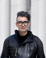 Person mit dunklem Haar und Brille trägt eine schwarze Lederjacke. Hintergrund ist eine helle Wand.