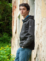 Eine Person lehnt an einer verfallenen Steinmauer im Grünen. Sie trägt Jeans und eine dunkle Jacke.