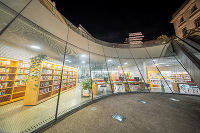 Moderne Bibliothek mit viel Glas, heller Innenraum mit Büchern und Sitzgelegenheiten. Nachtaufnahme mit Spiegelungen der Umgebung im Glas.